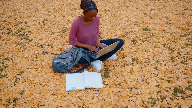 A young Black woman studying outdoors, on a leaf-covered lawn with a laptop computer balanced on her legs.
